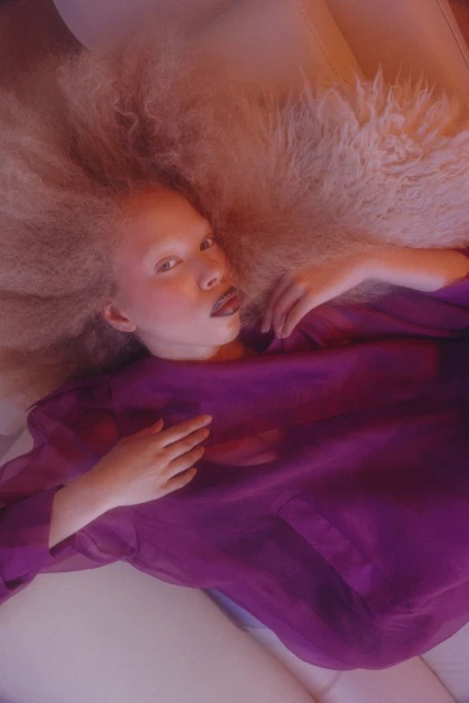 Portrait of a light-skinned person with large blonde hair lying on a cream-colored couch wearing a purple top in a brightly lit room.
