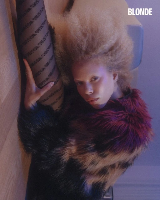 Albino woman lying on the floor wearing black Wolford tights, a multi-colored fur coat, and light blonde hair.