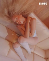 High-angle shot of a woman with voluminous blonde afro hair reclining on a cream modular sofa for BLONDE Magazine.