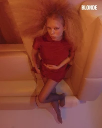 Model with voluminous frizzy hair wearing a red mini dress and dark sheer tights reclines on a high-back cream booth in warm light.
