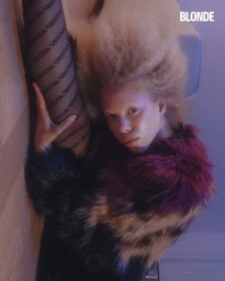 Model with blonde afro hair in a multi-toned faux fur coat and branded Wolford tights poses with one leg raised indoors.