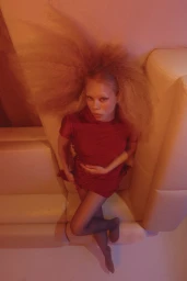 Woman in red dress with voluminous blonde hair seated against white sofa cushions in warm lighting