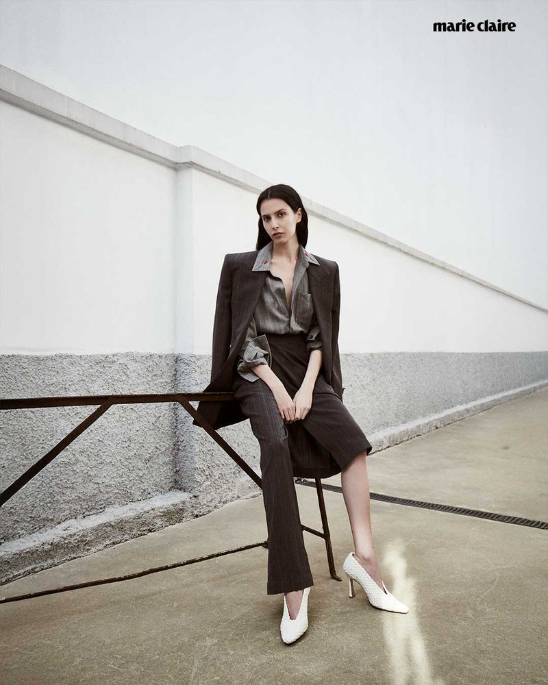 A woman in a gray blazer and skirt sits on a metal railing against a white wall in an outdoor fashion shot with white heels.