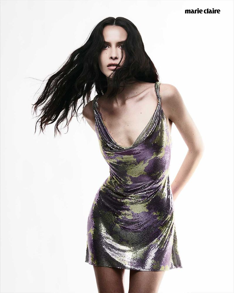 Portrait of a woman with dark hair, wearing a patterned mini dress, against a white background in a fashion editorial style.