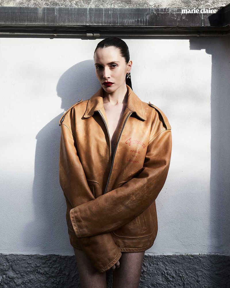 Portrait of a young woman wearing an oversized leather jacket, set against a textured white wall with magazine logo in the corner.