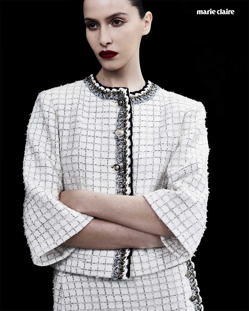 Elegant woman in a white tweed suit with arms crossed against a black background, fashion editorial in a studio with magazine branding.