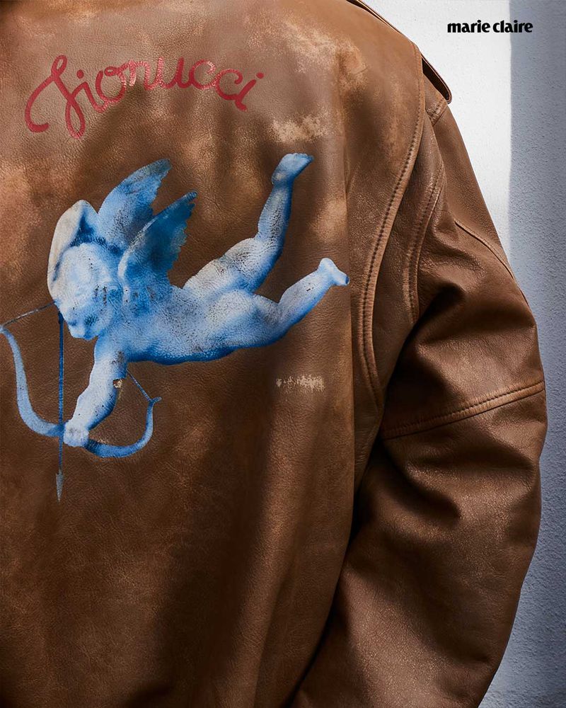 Brown leather jacket with a blue angel design and 'Fiorucci' text, set against a neutral background, Marie Claire logo in upper right.