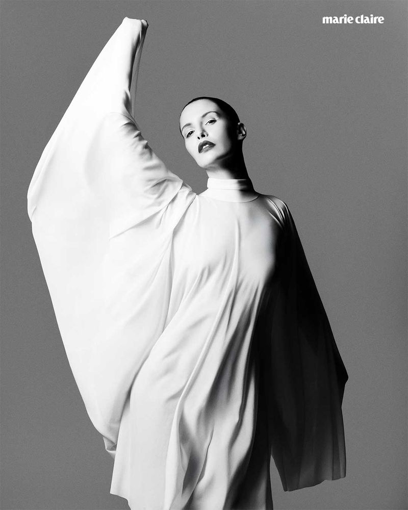 Black and white fashion portrait of an adult woman in a flowing white gown with raised arm, against a simple grey background.