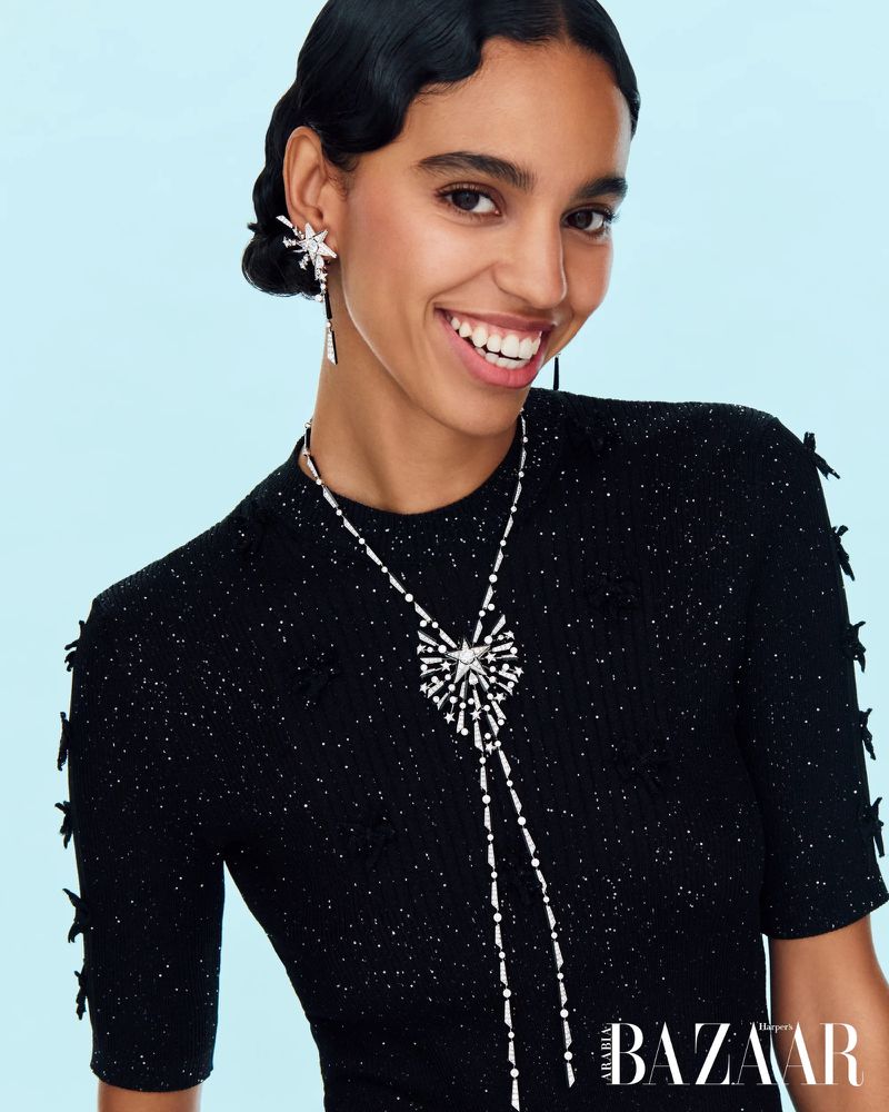 Woman wearing a black short-sleeved top and sparkly jewelry, including earrings and a star-shaped necklace, smiles against a light blue backdrop.