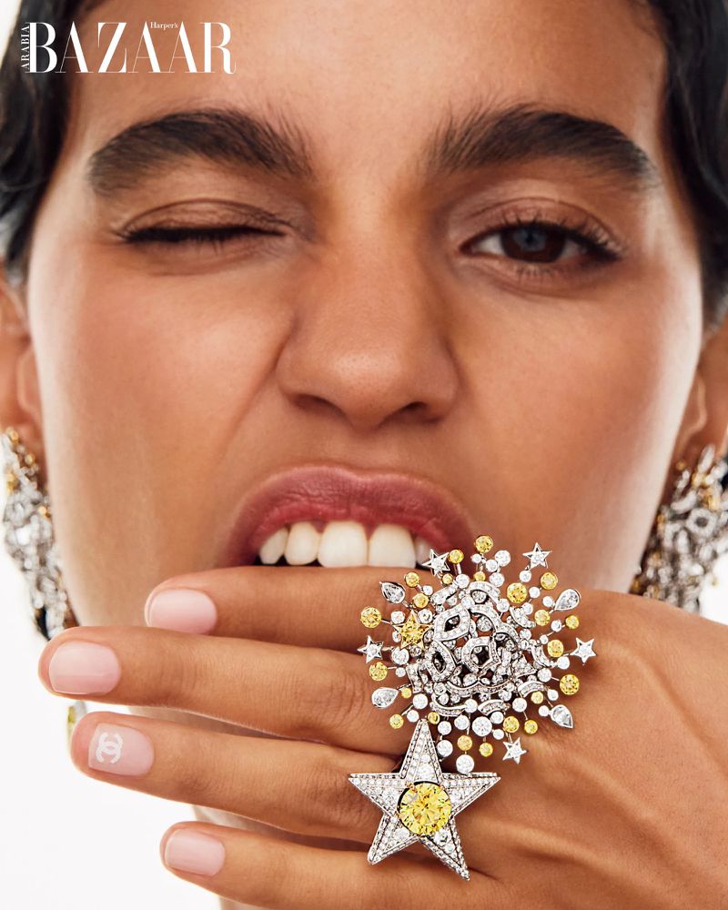 Close-up of a woman with fair skin winking, wearing large jeweled ring and earrings, posing in a studio setting; Bazaar magazine logo visible.