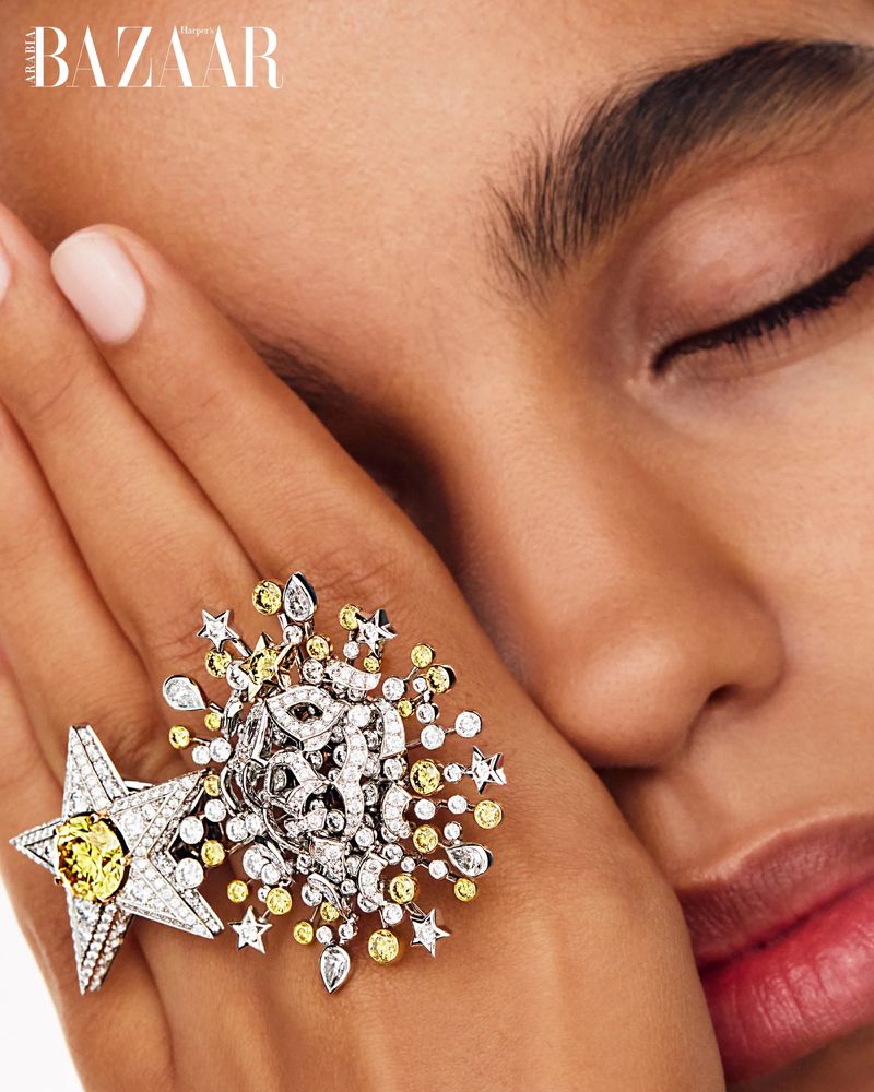 Close-up shot of a woman's face with eyes closed, adorned with a large decorative starburst ring featuring diamonds and yellow stones.