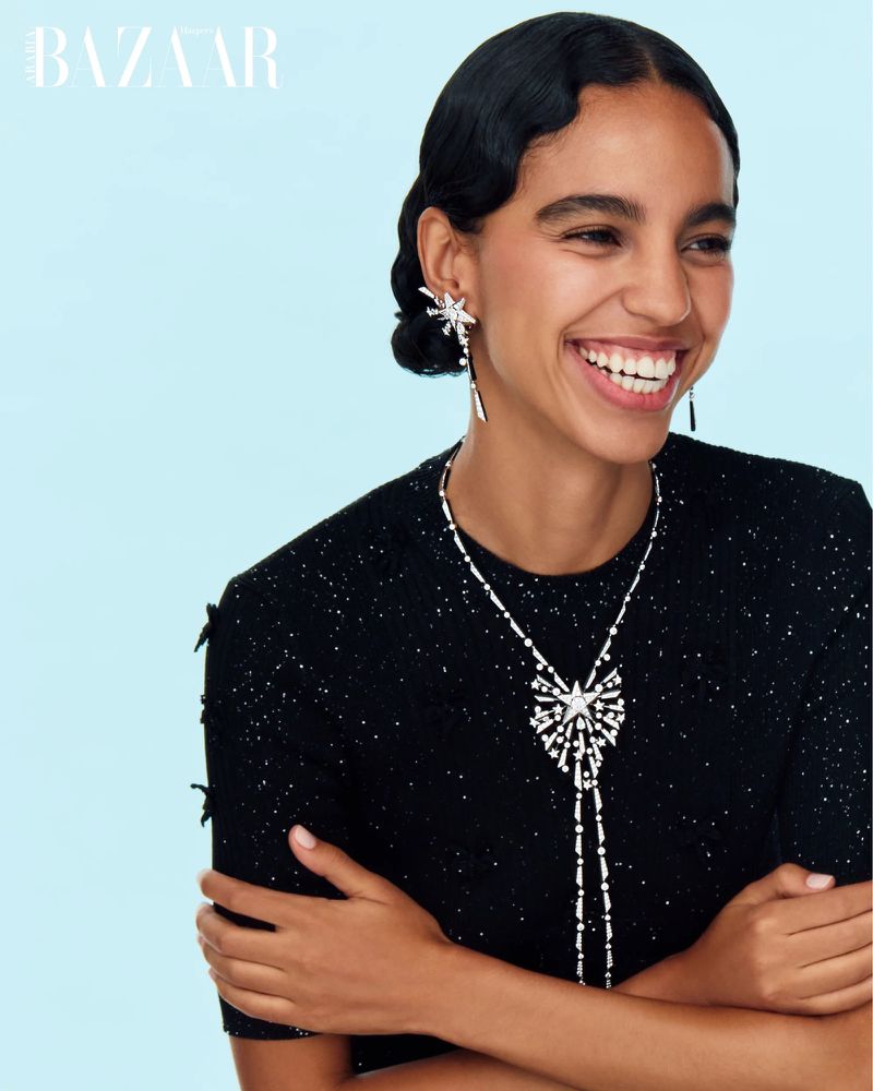 Close-up of a smiling woman in a black top with silver jewelry, including a star pendant, against a light blue studio backdrop.