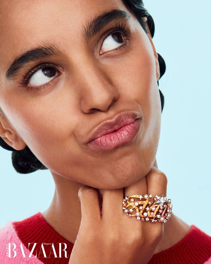 Close-up portrait of model Aouatif Saadi wearing a star-shaped gold and diamond Chanel ring under her chin on a light blue background.