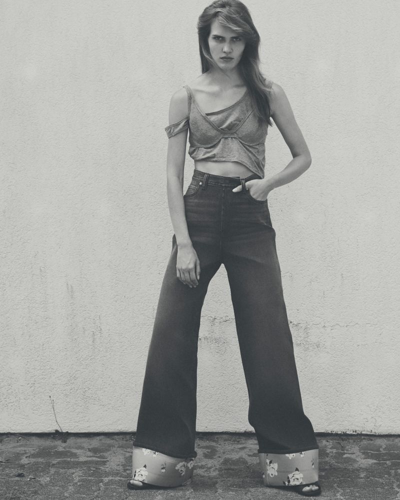 A stylish woman in a crop top and wide-leg jeans poses against a wall in a black-and-white fashion shot with vintage flair and bold design.