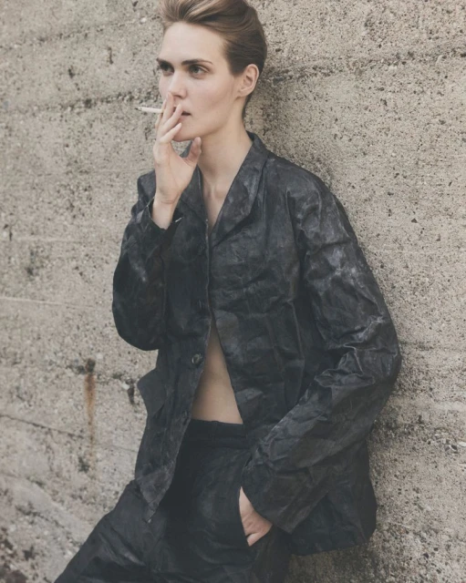 Adult female wearing a jacket and pants smoking a cigarette while leaning against a concrete wall in an outdoor setting.