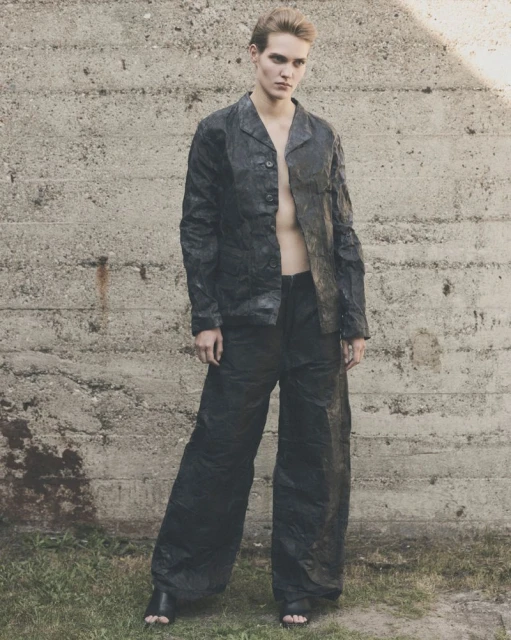A person posing in a dark textured jacket and wide leg pants against a weathered concrete wall in an outdoor setting with green grass.