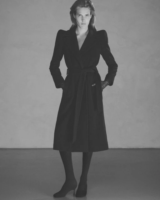 Black and white portrait of a female fashion model in a dark belted coat, dark hosiery, and high-heeled shoes, posed in a studio.