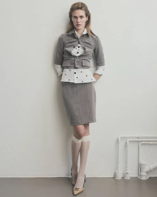 A fair-skinned woman poses in a gray skirt suit with a polka dot blouse indoors against a white wall, with visible pipes and tan pumps.