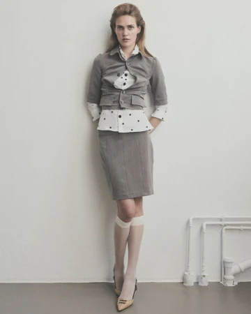 A fair-skinned woman poses in a gray skirt suit with a polka dot blouse indoors against a white wall, with visible pipes and tan pumps.