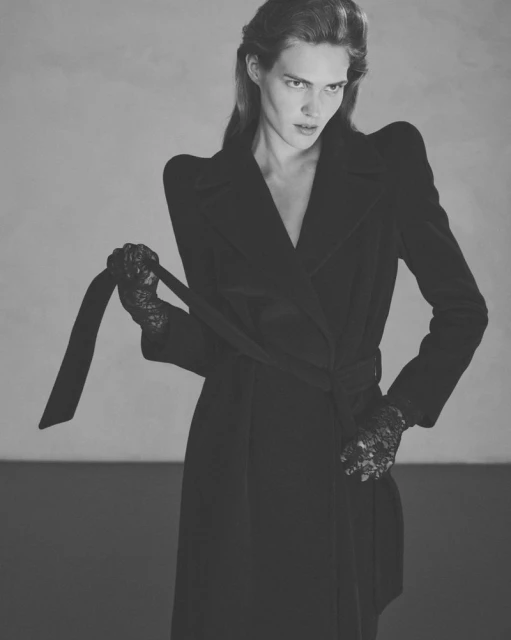 A woman in a black coat ties the belt with lace gloves in a black and white indoor studio fashion portrait, minimalist lighting.