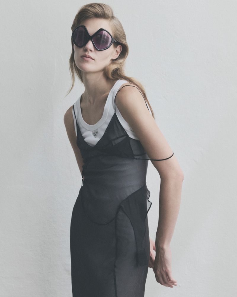 Woman wearing oversized dark sunglasses, a white tank top, and a layered sheer black dress against a plain pale background.