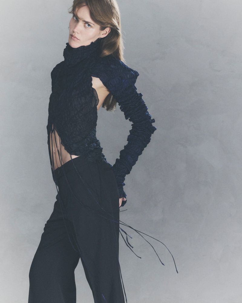 Model posing in an avant-garde black textured top with ruched long sleeves and black trousers against a textured grey backdrop.