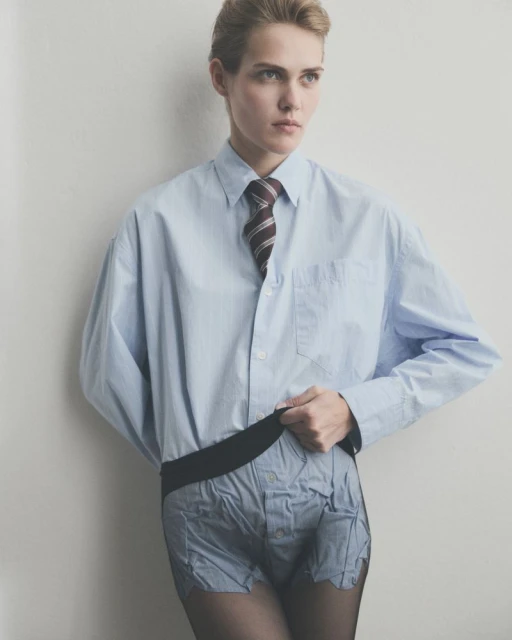 Adult female fashion model poses indoors wearing a light blue collared shirt, striped tie, repurposed dress-shirt shorts, and black tights.