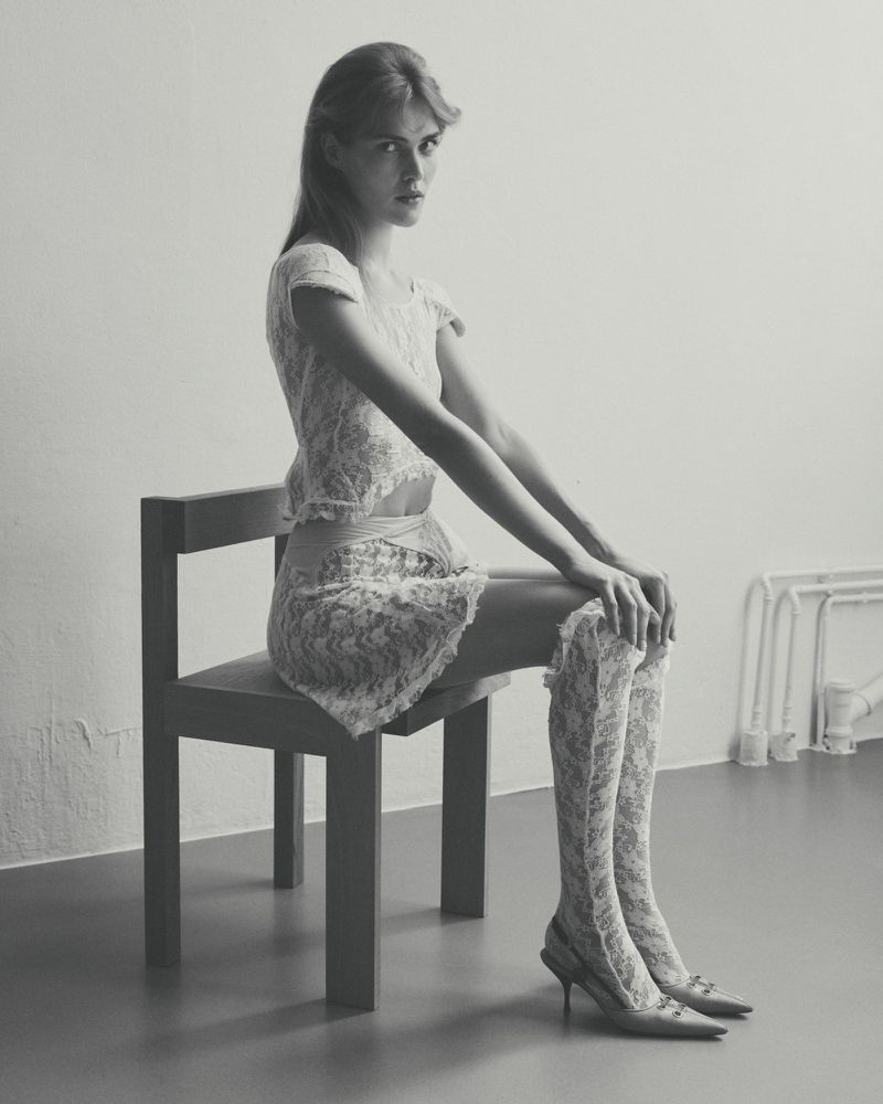 A young woman with long hair, wearing a lace top, skirt, thigh-high socks and heels, sits on a chair in a minimalist studio setting.