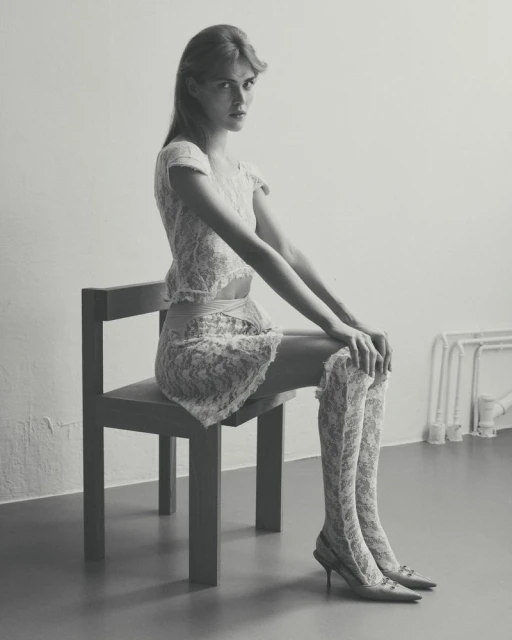 A young woman with long hair, wearing a lace top, skirt, thigh-high socks and heels, sits on a chair in a minimalist studio setting.