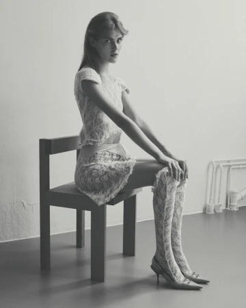 A young woman with long hair, wearing a lace top, skirt, thigh-high socks and heels, sits on a chair in a minimalist studio setting.