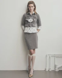 Model standing against a white wall wearing a grey jacket, skirt, and polka dot shirt with knee-high socks.