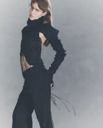 Model posing in an avant-garde black textured top with ruched long sleeves and black trousers against a textured grey backdrop.