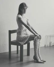 Model seated on a wooden chair wearing a white lace matching top and skirt with knee-high lace socks and heels.