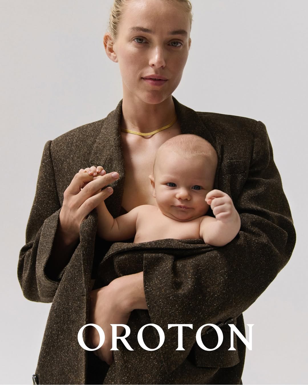Fashion shot of a mother holding her baby, both in a studio setting, showcasing a brown blazer and a delicate gold necklace.