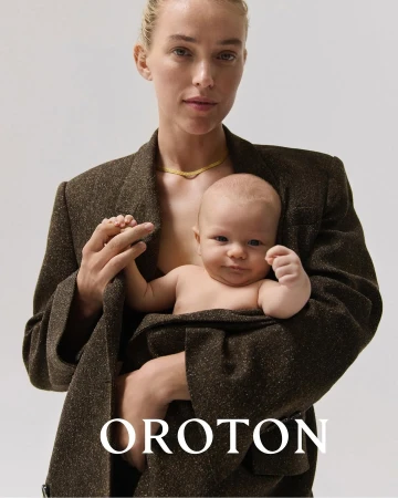 Fashion shot of a mother holding her baby, both in a studio setting, showcasing a brown blazer and a delicate gold necklace.