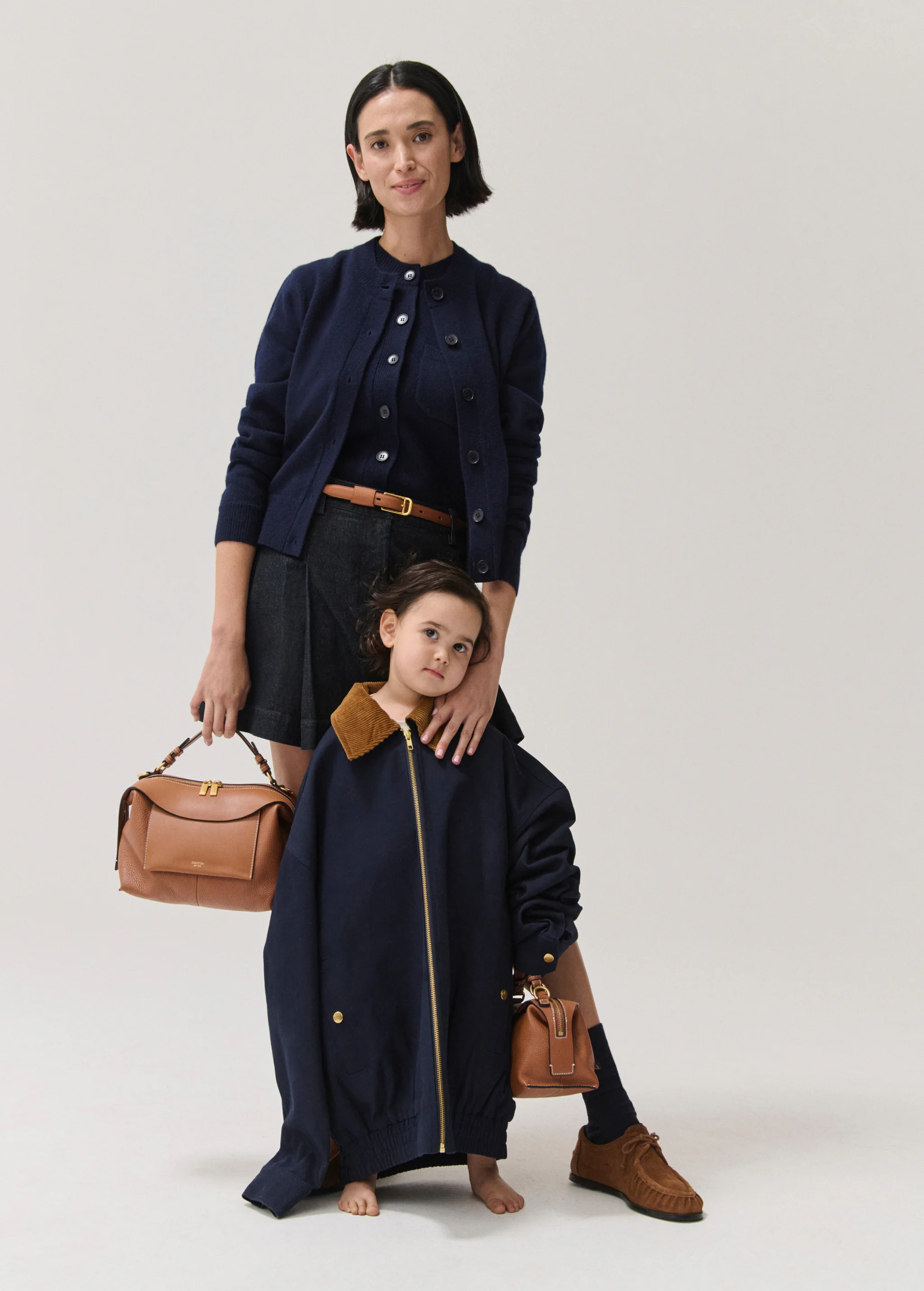 Woman and child in studio. Woman wears navy cardigan and skirt with a brown belt and is holding a purse. Child wears oversized jacket.