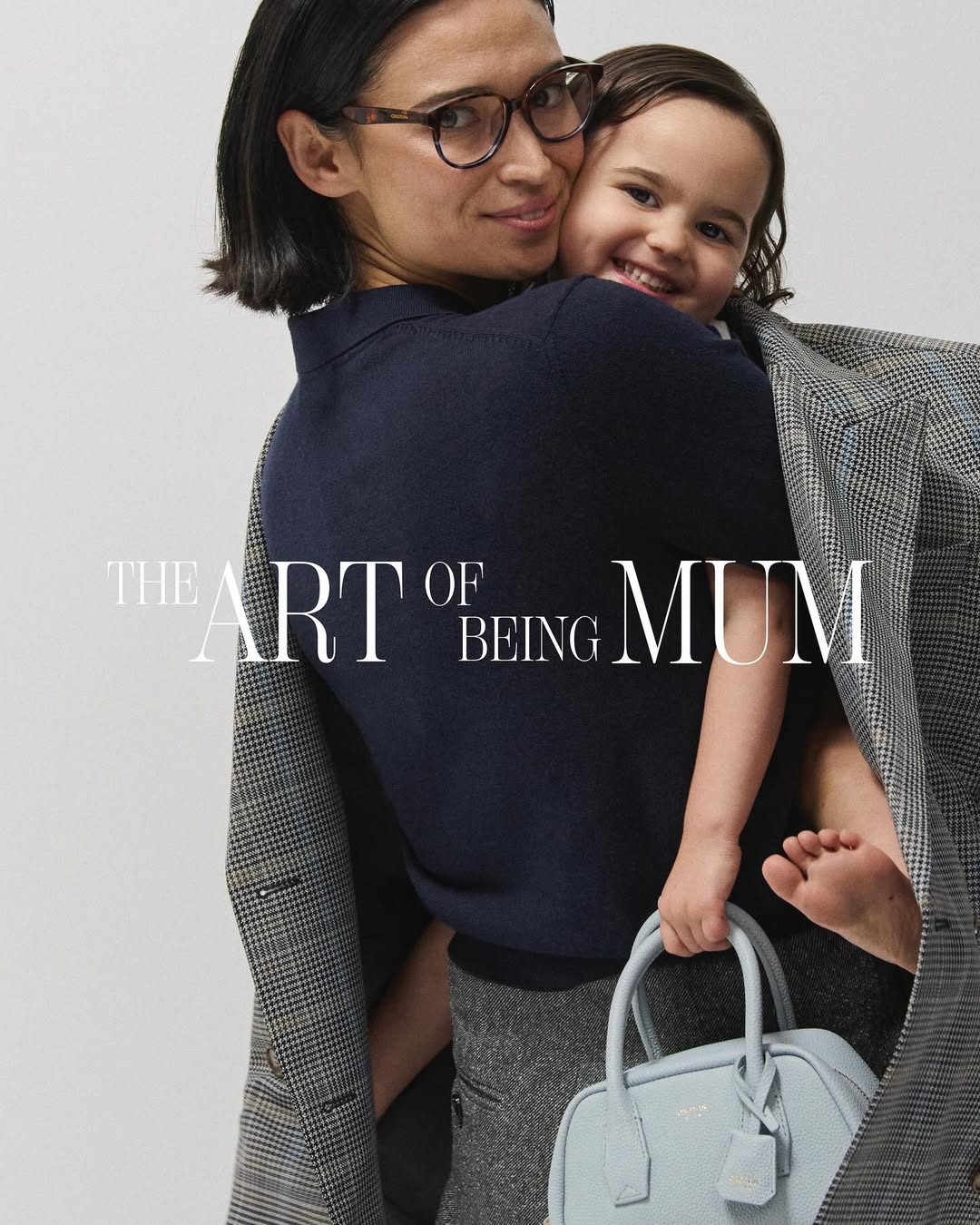Indoor portrait shot of an adult woman wearing glasses holding a young child with a light blue purse in a simple studio environment.