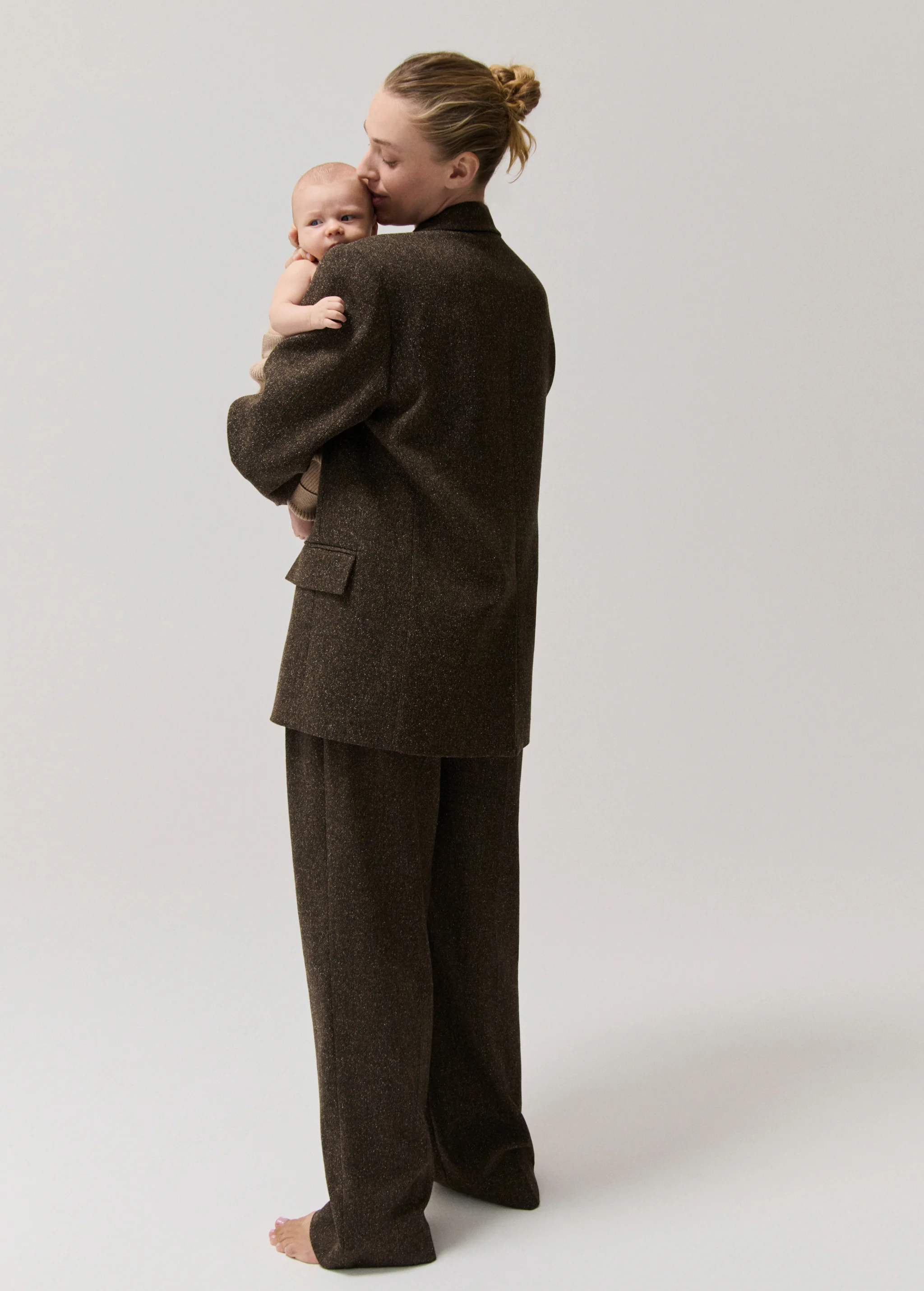 A woman in a brown suit holds a baby against a light gray background. The woman kisses the baby on the forehead.