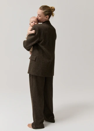 A woman in a brown suit holds a baby against a light gray background. The woman kisses the baby on the forehead.