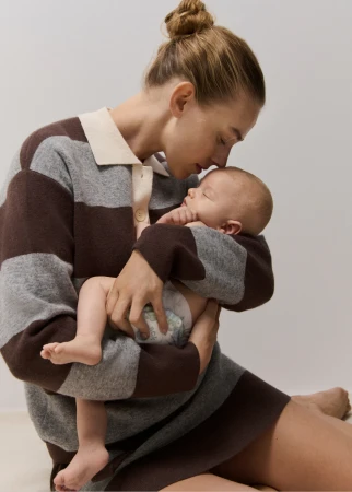 A mother wearing a brown and grey striped sweater dress holds a baby in her arms, set against a neutral backdrop in a lifestyle shot.
