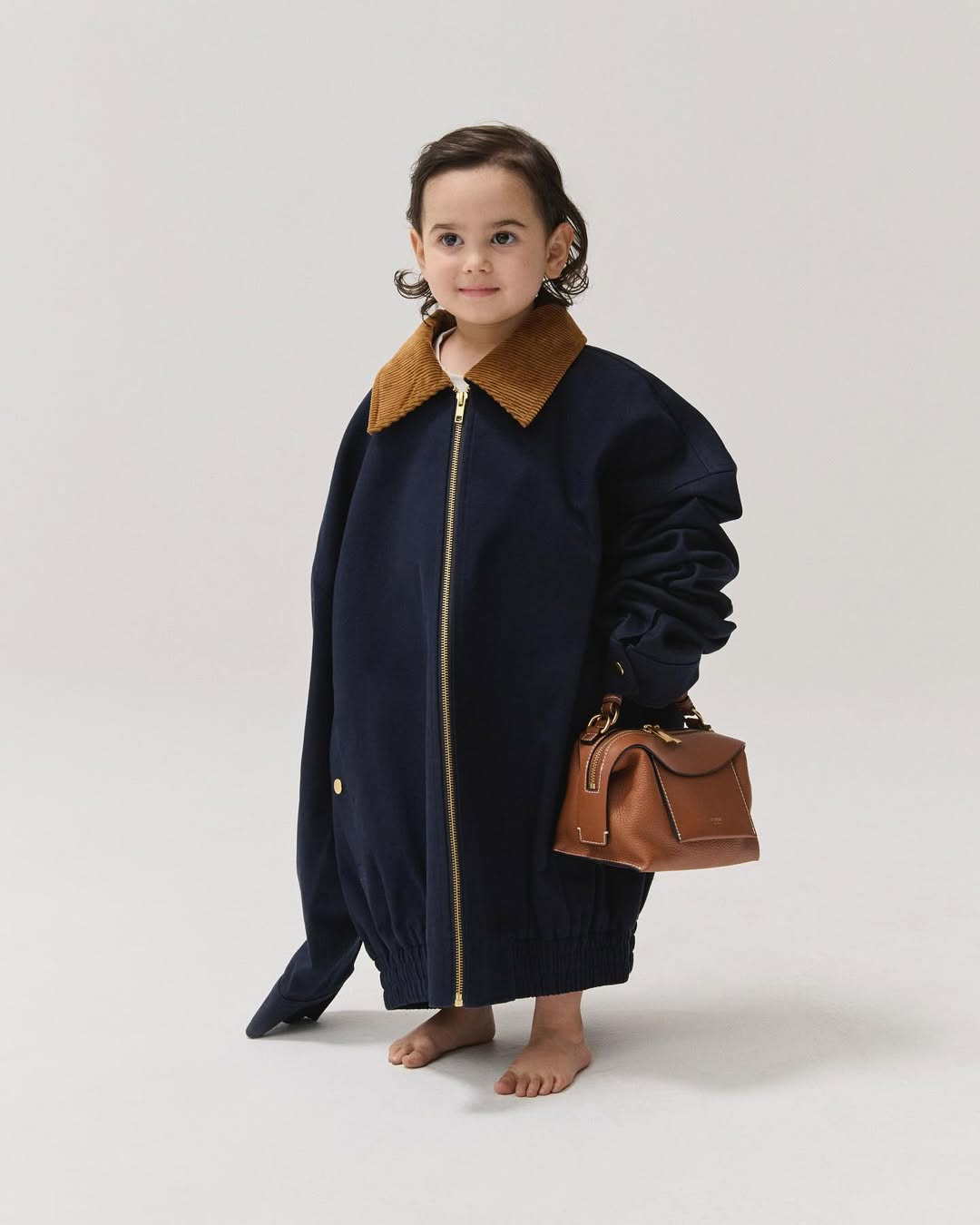 A young child models an oversized navy blue corduroy jacket with a brown leather handbag against a plain white background indoors.