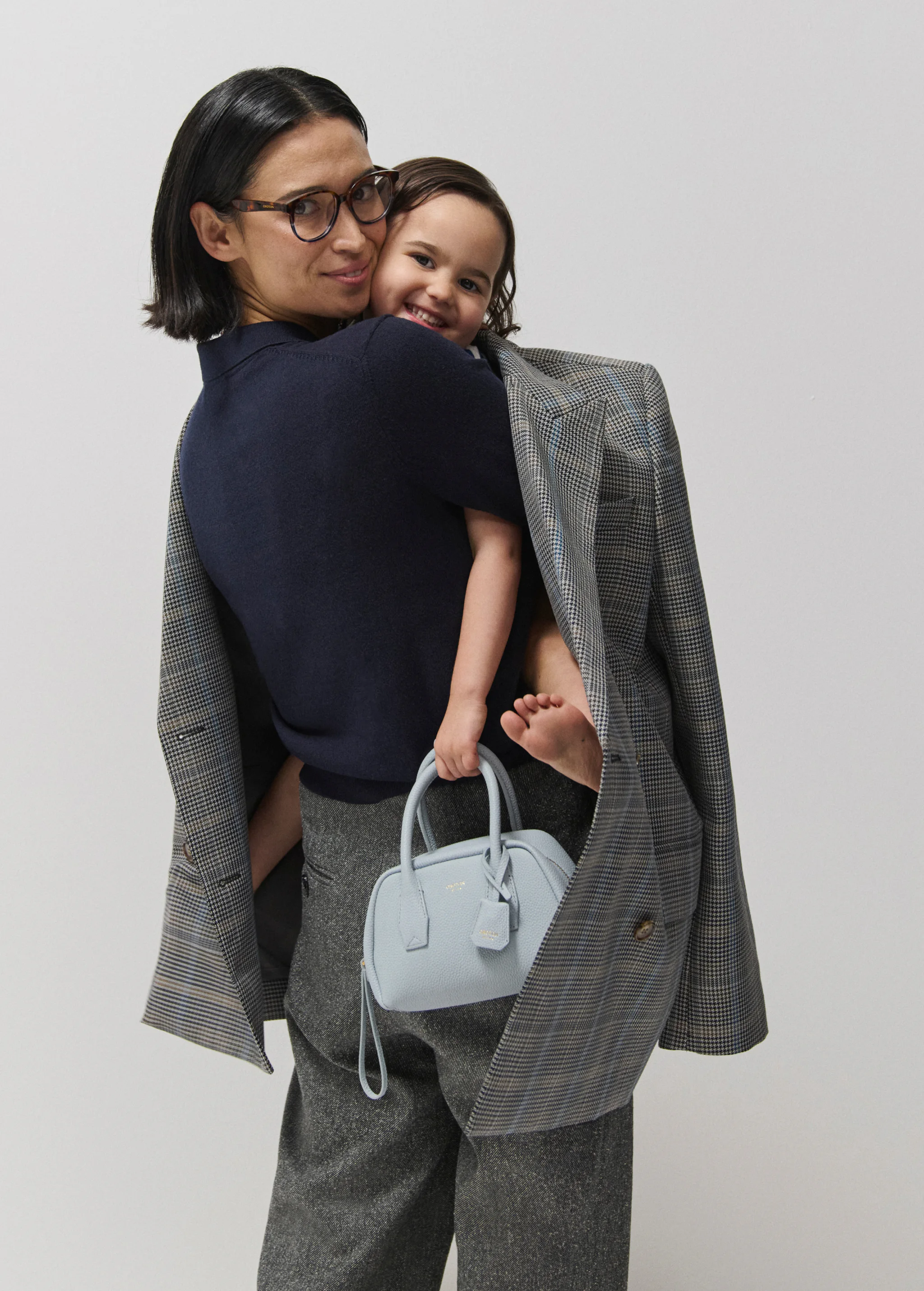 Adult female carries a toddler, holds a blue handbag, wears a grey blazer and pants on a plain studio background. Minimalist fashion.
