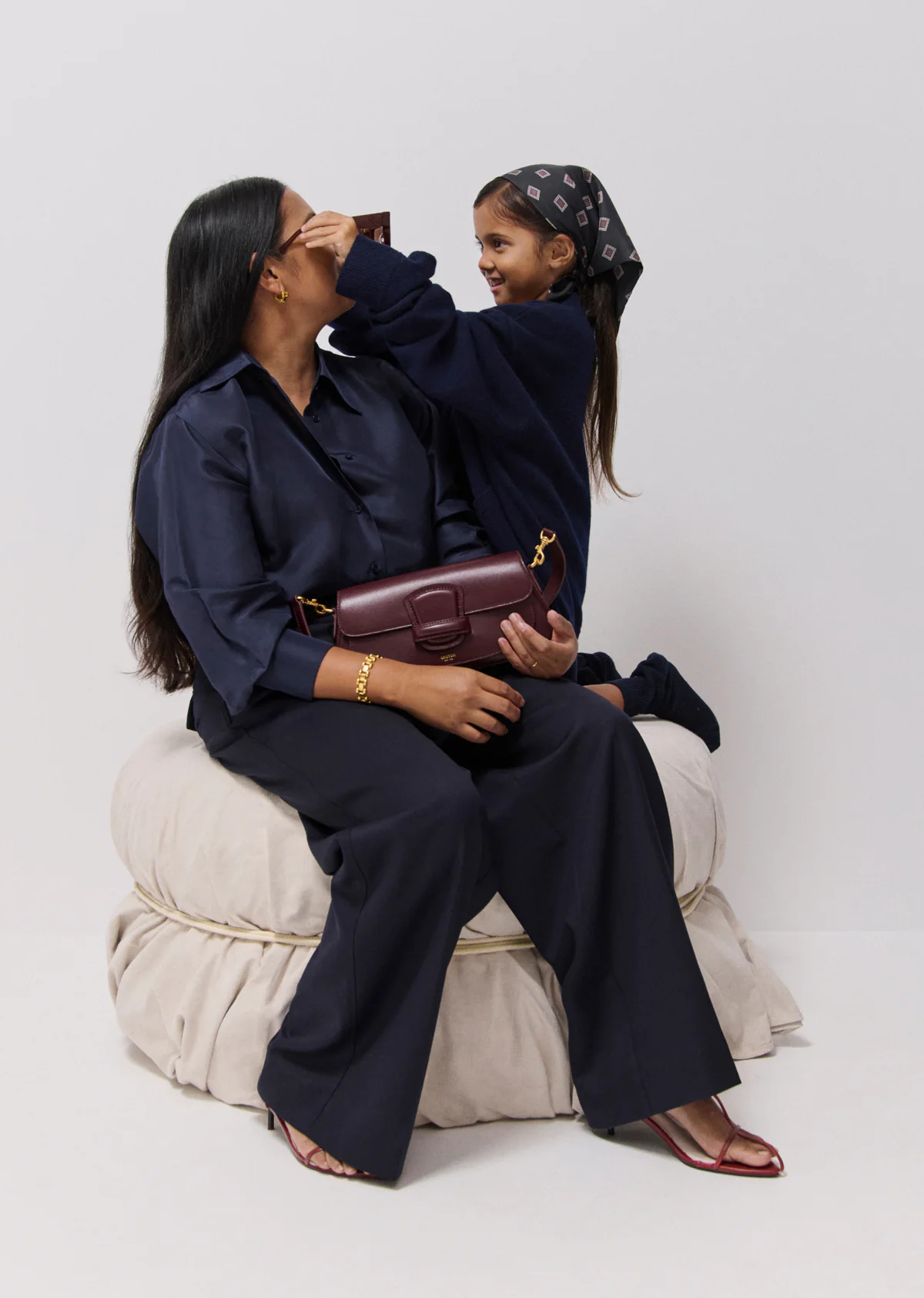 A woman and child pose in an indoor setting, showcasing a dark red leather handbag, both are in navy blue clothing.