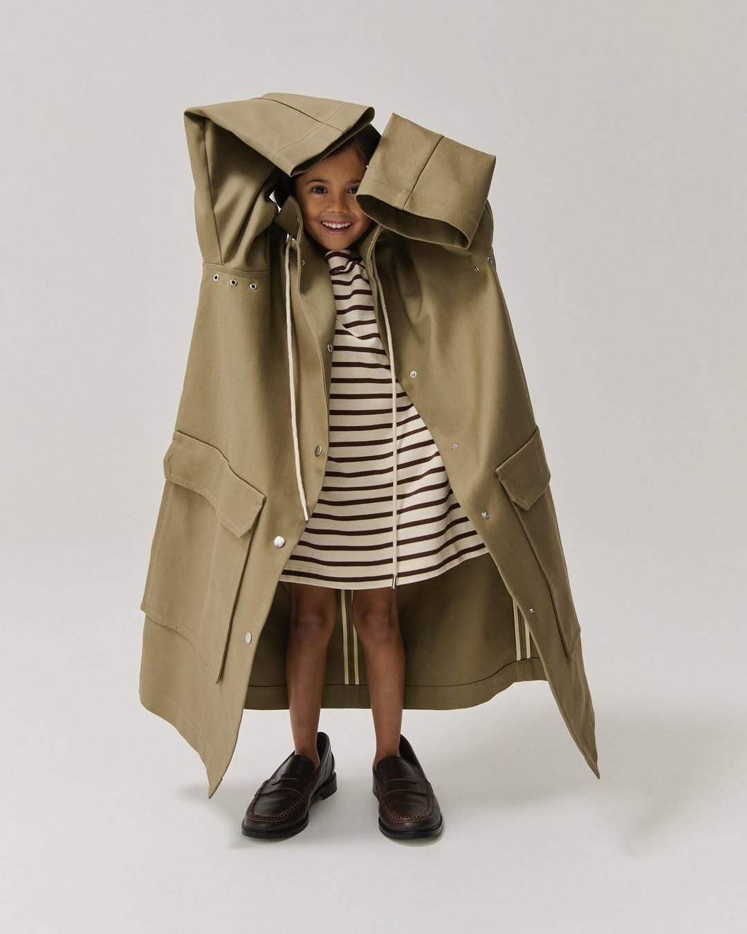 A cute child model poses in a beige striped dress and oversized trench coat with brown penny loafers against a neutral backdrop.