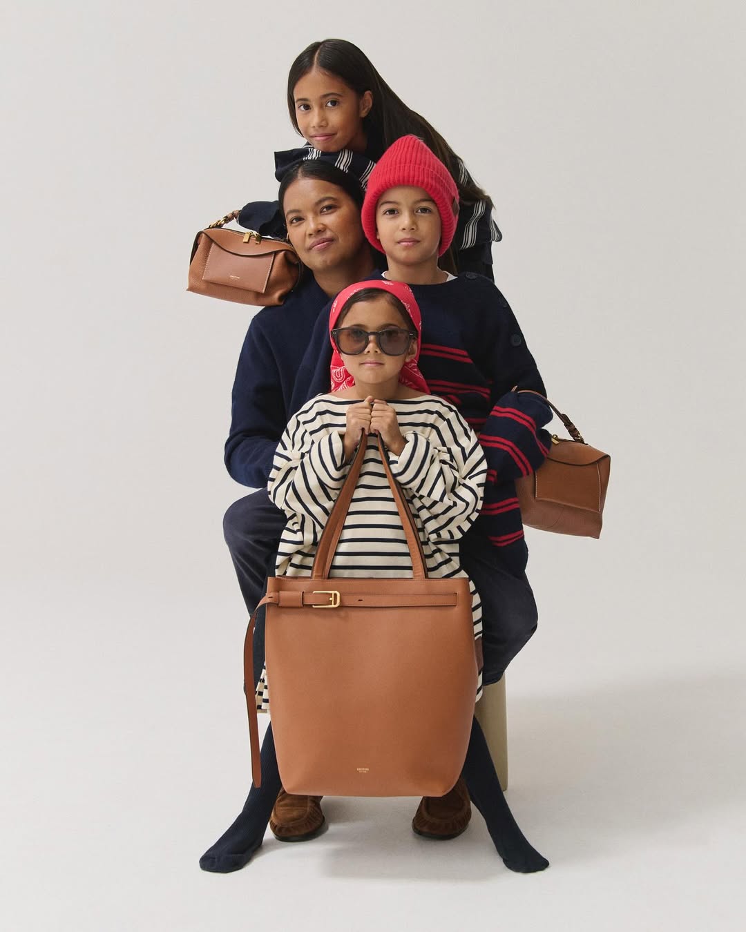 A family of four poses in a photo studio with light skin, wearing fashionable clothing and displaying brown leather handbags against a white backdrop.