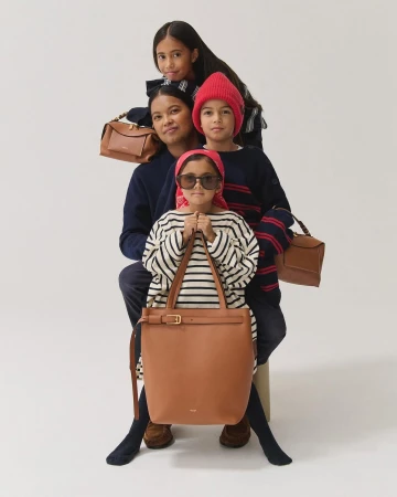 A family of four poses in a photo studio with light skin, wearing fashionable clothing and displaying brown leather handbags against a white backdrop.