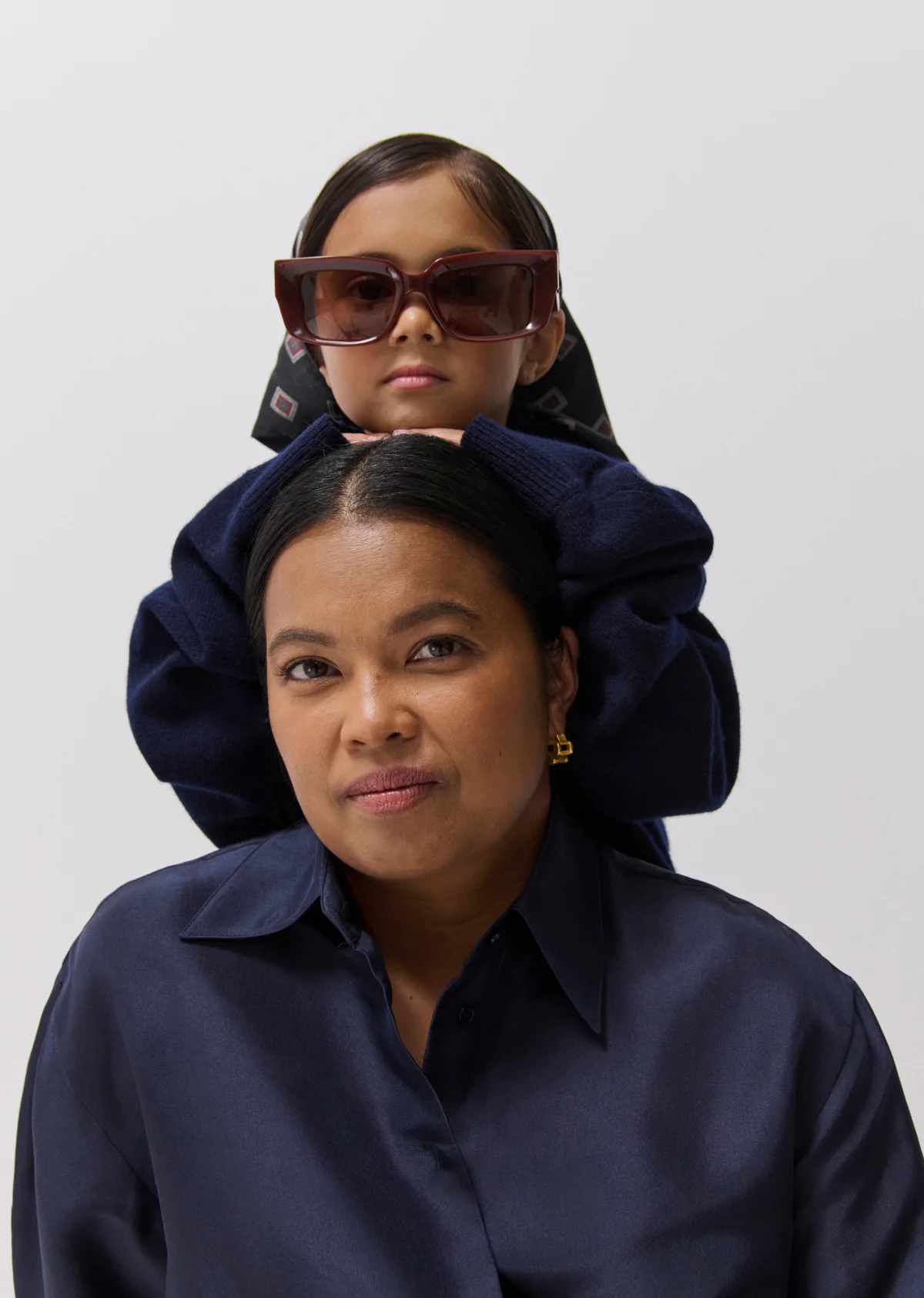 Mother and child portrait in indoor studio. The child wears oversized sunglasses and sits atop the mother's shoulders, smiling.