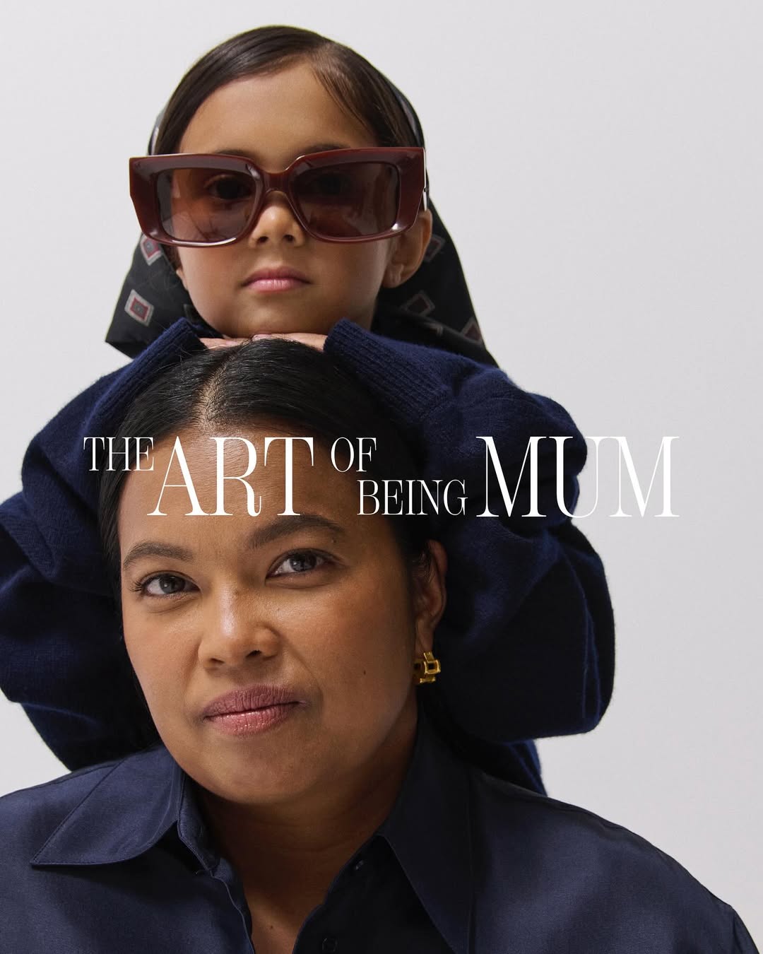Mother and daughter portrait in photo studio. Child sits on woman's shoulders wearing oversized sunglasses in a family moment.
