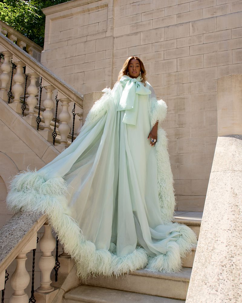 Elegant woman in a light green gown with feathered trim poses on stone stairs, showcasing a blend of fashion and architectural elements.