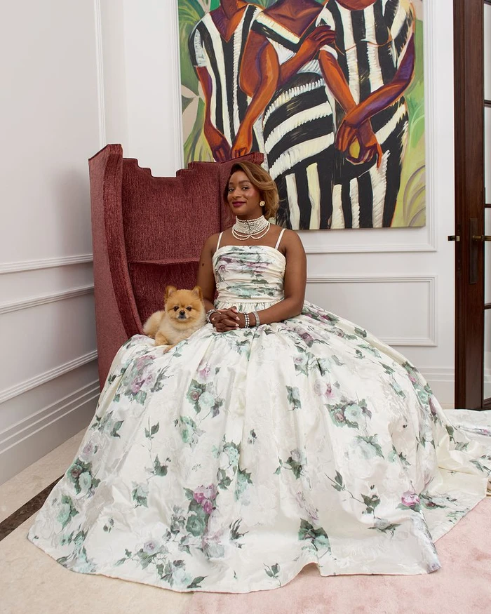Woman in floral ball gown seated in a burgundy wingback chair holding a small dog with an abstract painting behind her